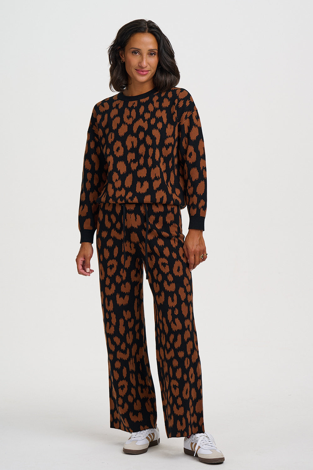 A woman modeling a matching two-piece lounge set featuring a brown and black leopard print pattern, consisting of a long-sleeve top and wide-leg pants, paired with white sneakers against a light gray background.