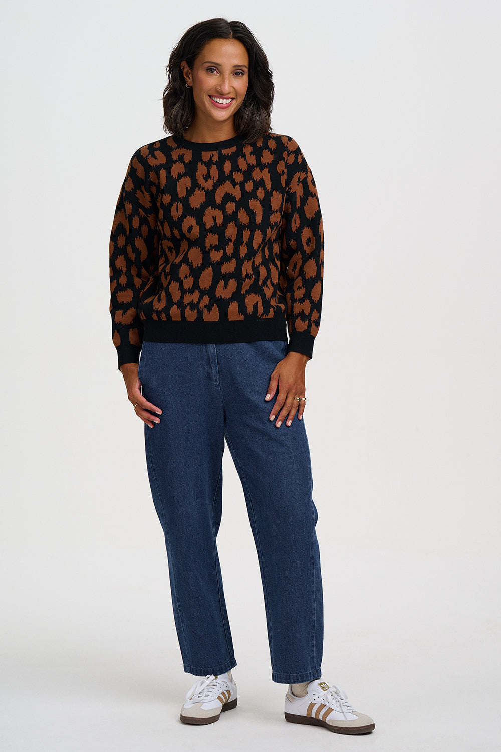 A woman modeling a black crewneck sweater with a brown leopard print pattern, paired with dark blue jeans and white sneakers, against a light gray studio background.