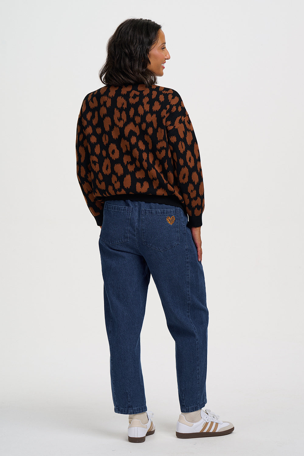 A model shown from the back wearing a black and brown leopard print sweater, blue jeans, and white sneakers against a light gray studio background.