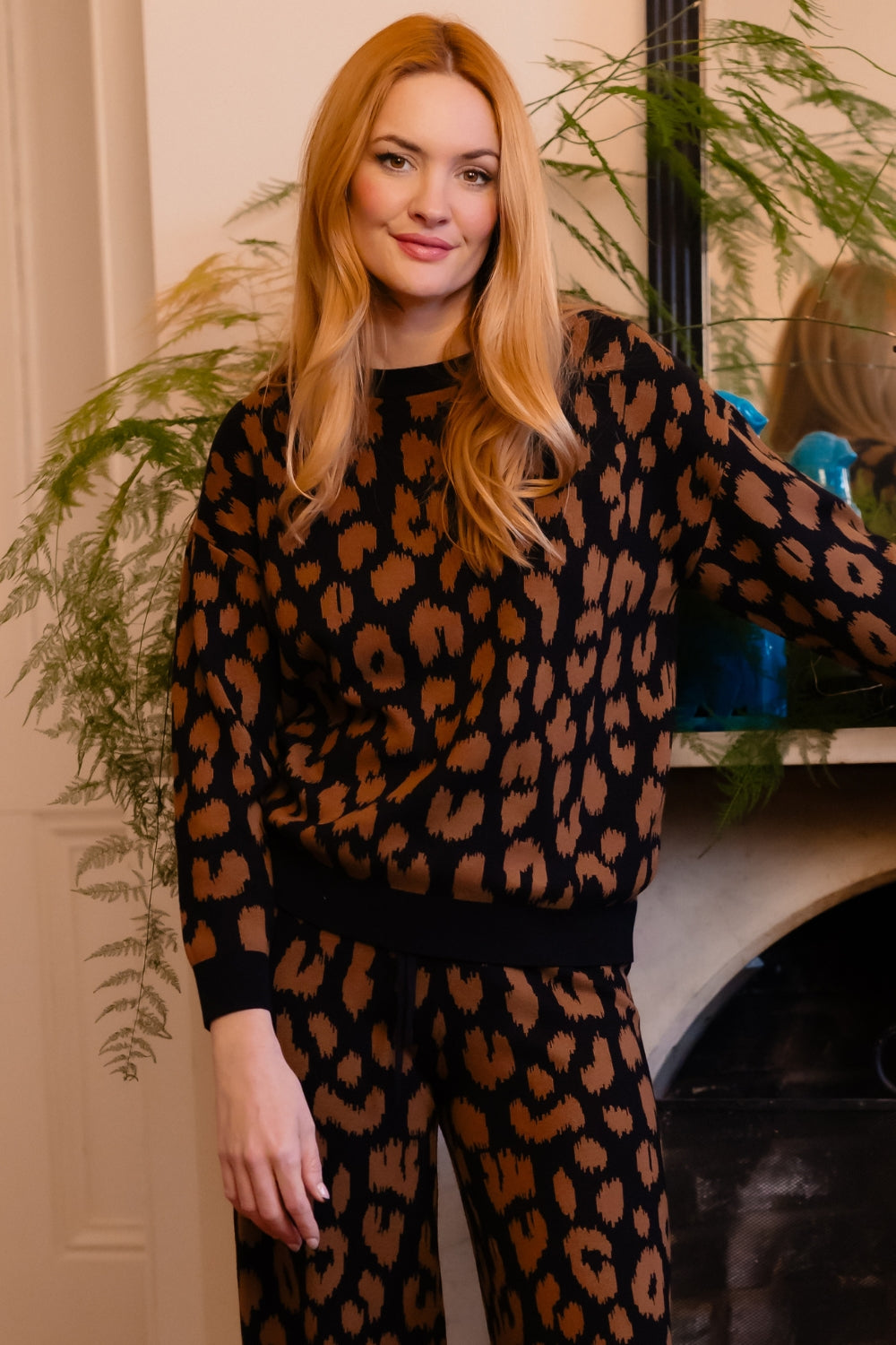 A woman with long blonde hair wearing a matching black and brown leopard print knit lounge set, standing in front of a fireplace with plants in the background.