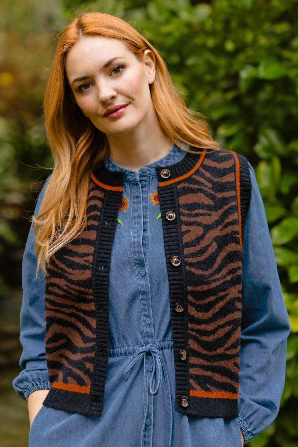 A woman with long red hair wearing a blue denim jumpsuit with a brown and black tiger-stripe knit vest over it, standing outdoors in front of a blurred green foliage background.