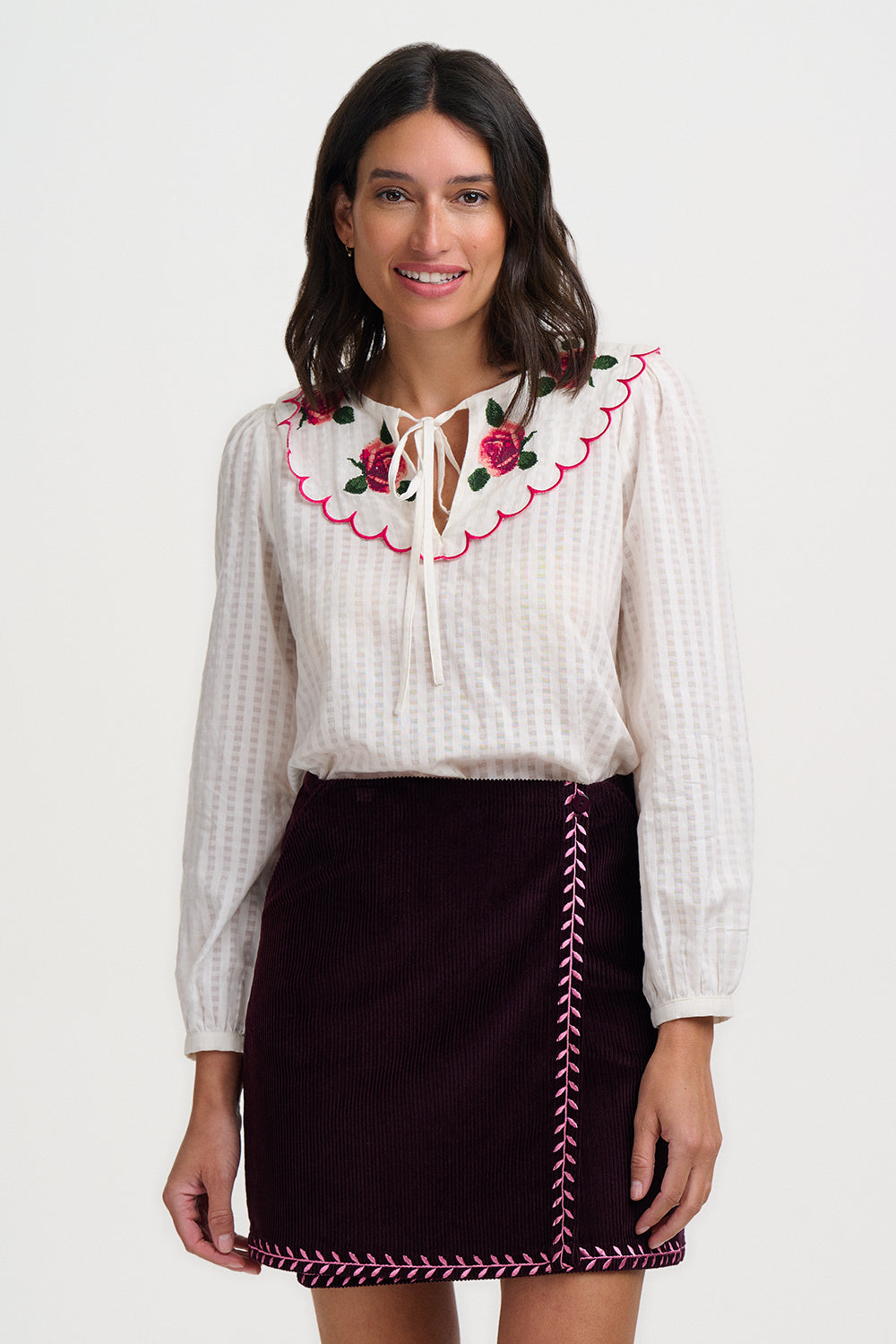 Woman wearing a white blouse with floral embroidery and a black skirt with red stitching on a white background