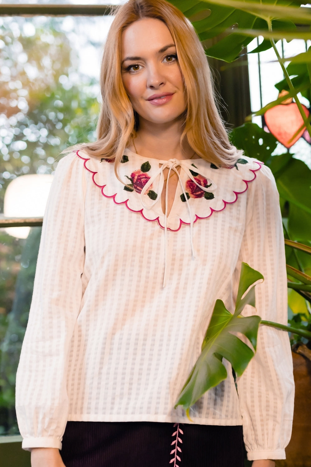 A woman wearing a cream-colored seersucker blouse with a scalloped Peter Pan collar decorated with pink and green floral embroidery posed in front of a window with green plants.
