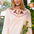 A woman wearing a cream-colored seersucker blouse with a scalloped Peter Pan collar decorated with pink and green floral embroidery posed in front of a window with green plants.