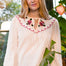 A woman wearing a cream-colored seersucker blouse with a scalloped Peter Pan collar decorated with pink and green floral embroidery posed in front of a window with green plants.