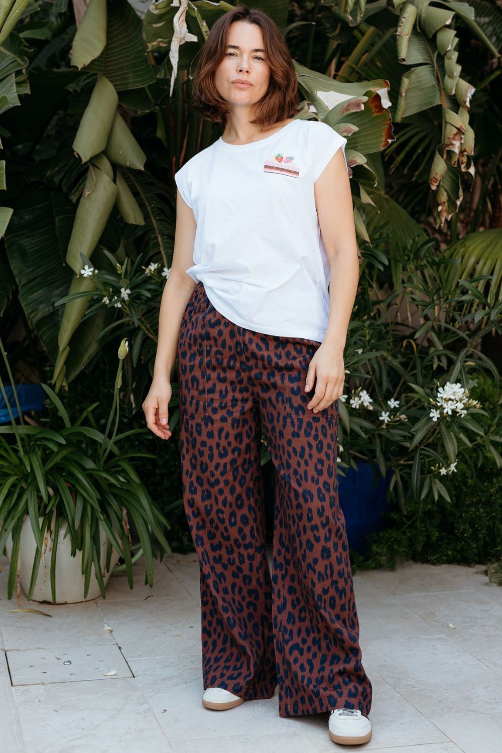 Woman wearing a white t-shirt and leopard print pants standing in front of green plants.
