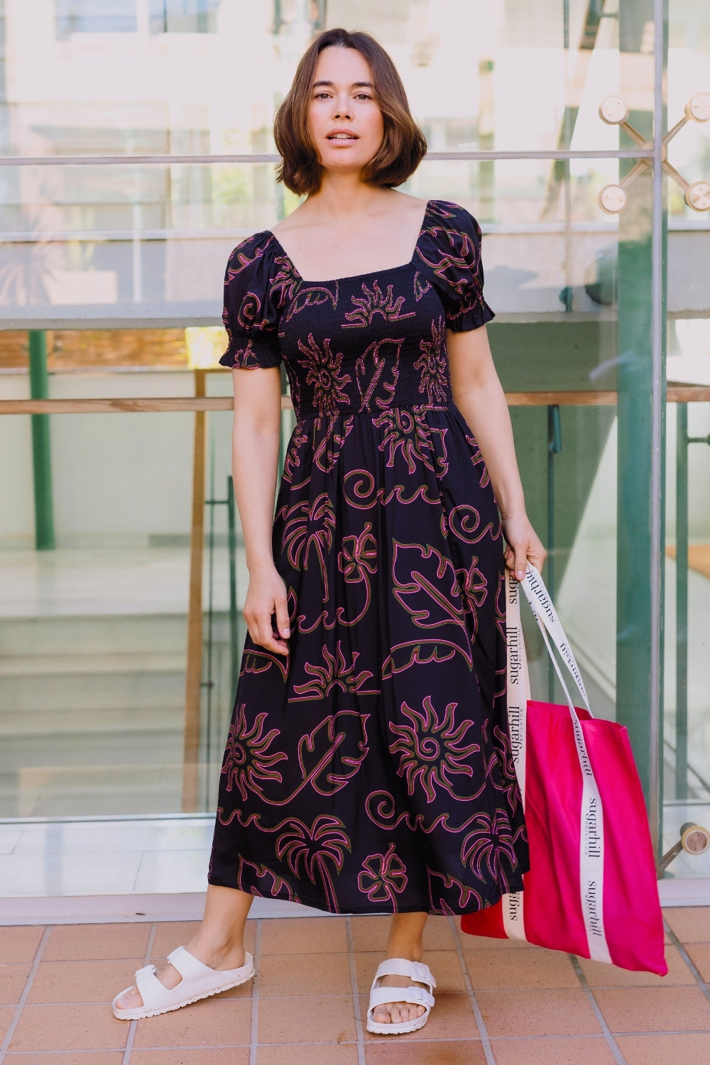 Woman in a patterned dress holding a pink bag outside a building.