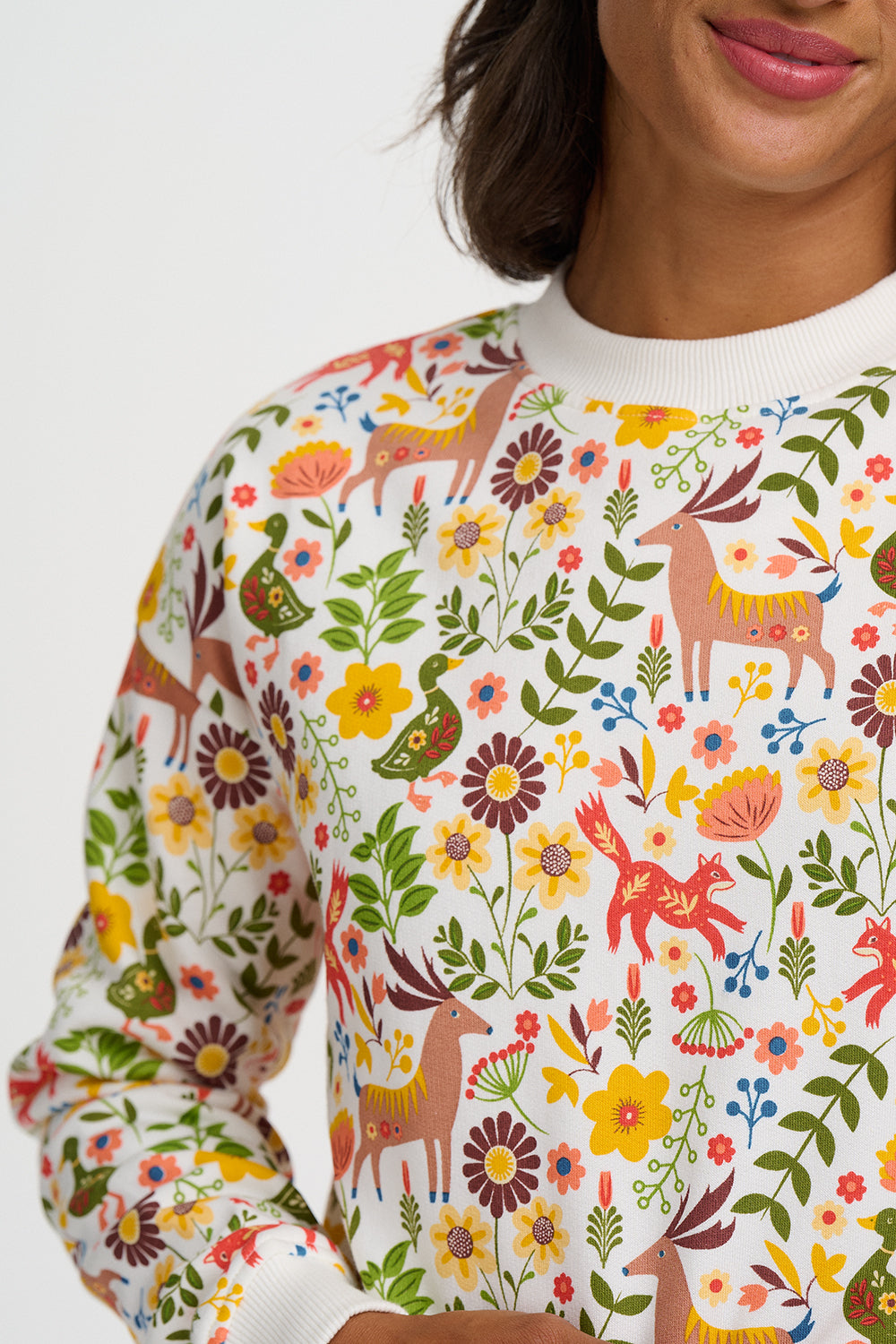 A close-up of a person wearing a white crewneck sweater covered in a colorful, whimsical forest pattern featuring deer, flowers, leaves, and birds.