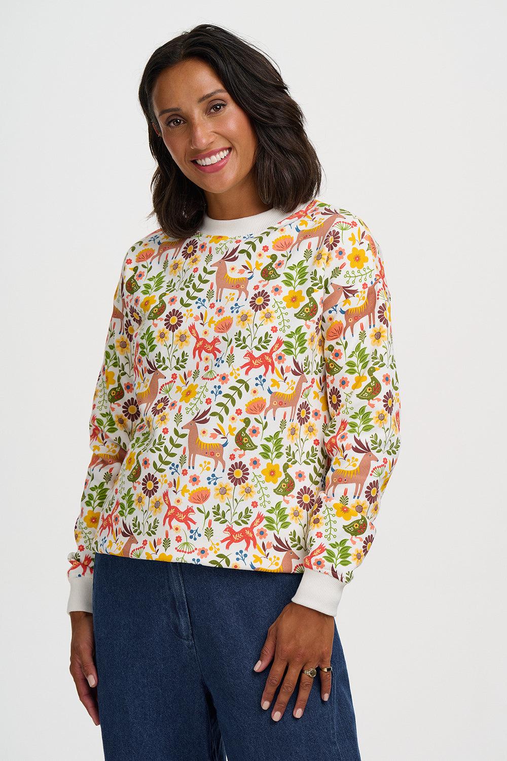 A woman wearing a long-sleeve crewneck sweatshirt with a colorful forest and deer pattern, paired with dark blue jeans, against a light gray studio background.