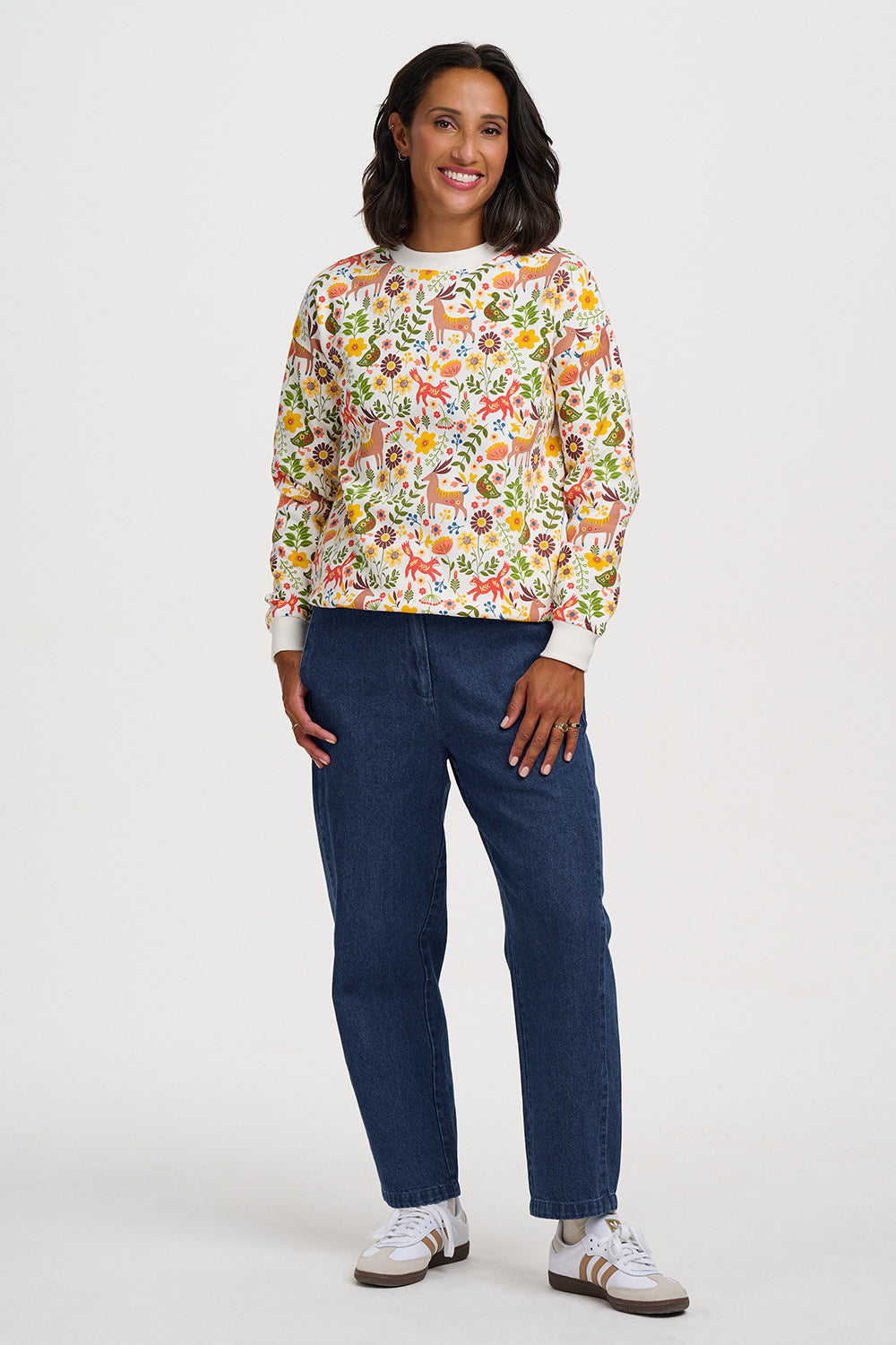 A woman modeling a long-sleeve crewneck sweatshirt with a dense multicolor floral and botanical pattern on a cream background, paired with dark blue jeans and white sneakers, against a plain light gray studio background.