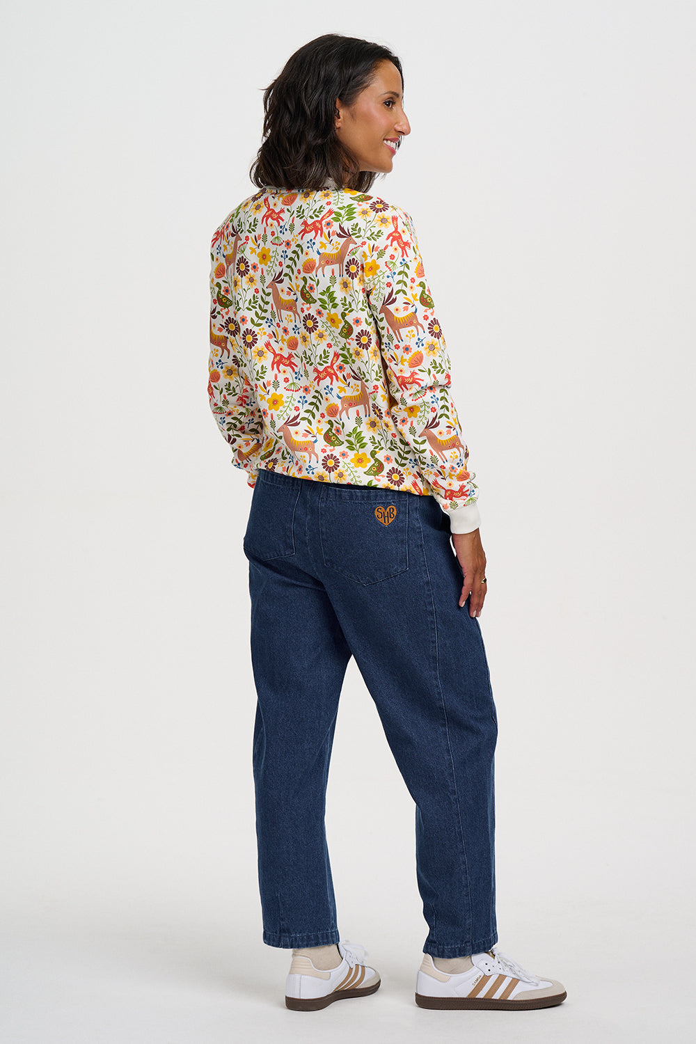 A model is shown from the back wearing a cream-colored long-sleeve bomber jacket with a colorful, whimsical animal and floral print, paired with dark blue jeans and white sneakers with gold stripes.
