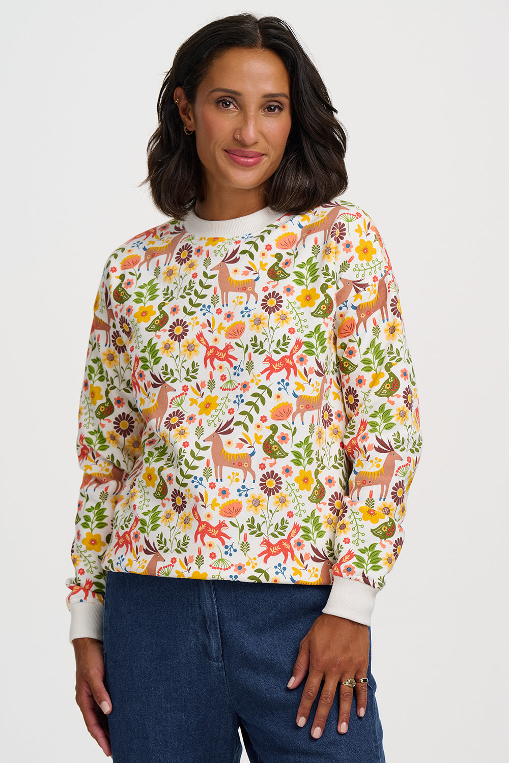 A woman wearing a long-sleeve crewneck sweatshirt with a colorful forest and deer pattern, paired with dark blue jeans, against a light gray studio background.