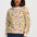 A woman wearing a long-sleeve crewneck sweatshirt with a colorful forest and deer pattern, paired with dark blue jeans, against a light gray studio background.