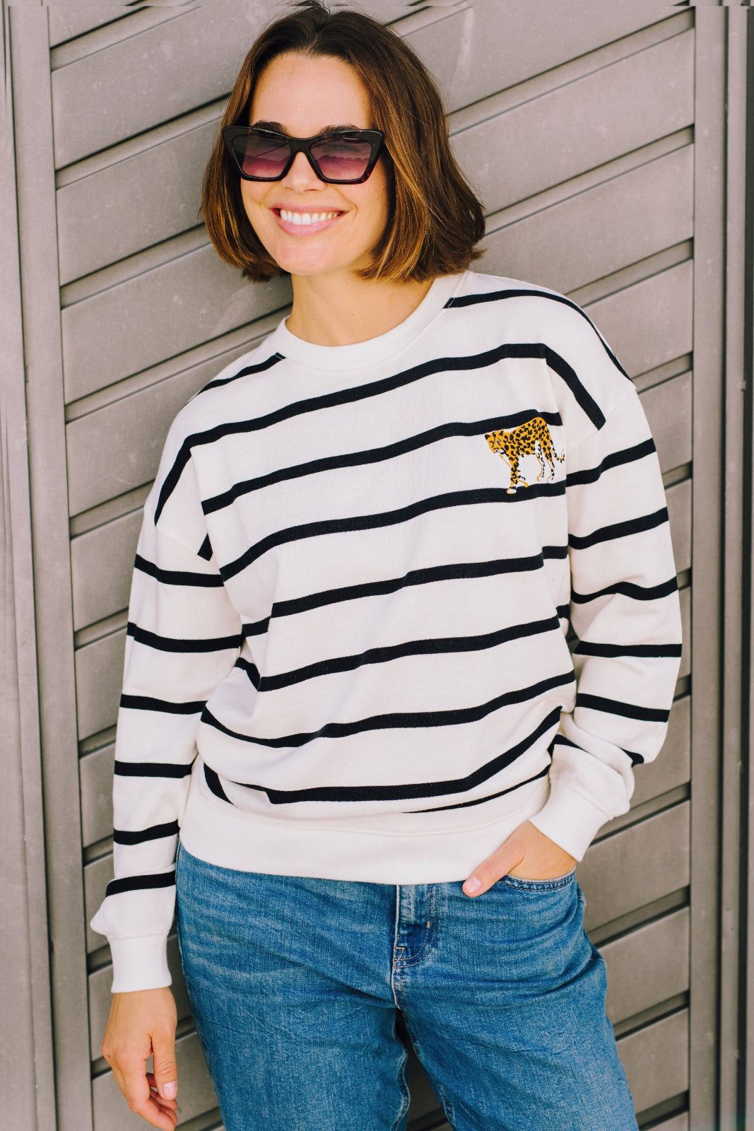 A woman wearing an off-white sweatshirt with black leopard embroidery, paired with blue jeans and sunglasses, standing in front of a wooden background.