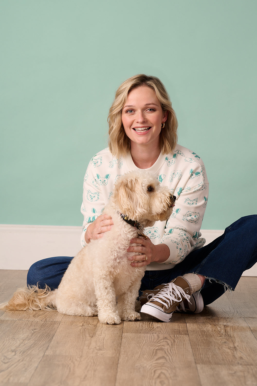 A person sitting on the floor wearing a sweatshirt with off-white and green colors, and a brown dog sitting on a blanket in front of them.