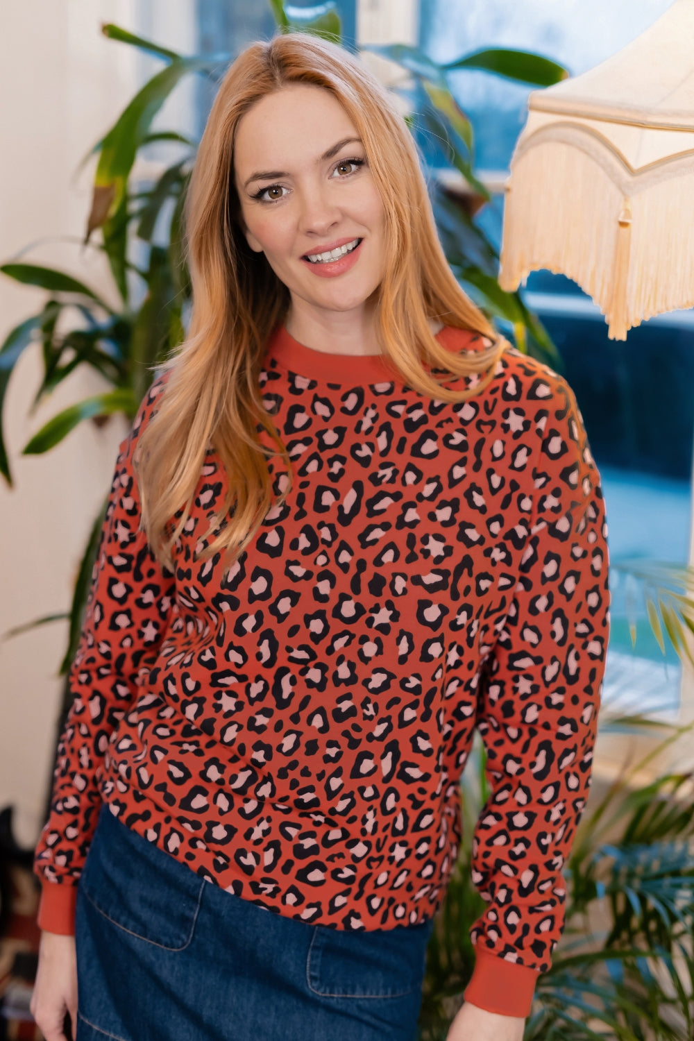 A woman with blonde hair wearing a rust-orange long-sleeve crewneck sweater with a black and white leopard print pattern, paired with a blue denim skirt. She is standing in an indoor setting with plants and a lamp in the background.