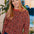 A woman with blonde hair wearing a rust-orange long-sleeve crewneck sweater with a black and white leopard print pattern, paired with a blue denim skirt. She is standing in an indoor setting with plants and a lamp in the background.