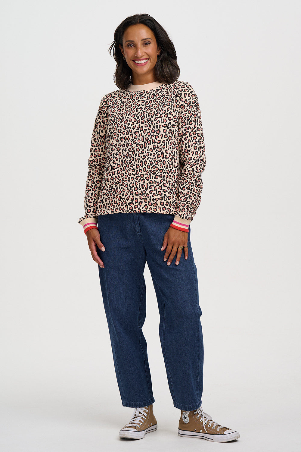 A woman modeling a long-sleeve crewneck top with a beige and brown leopard print, paired with dark blue jeans and brown sneakers, against a light gray studio background.