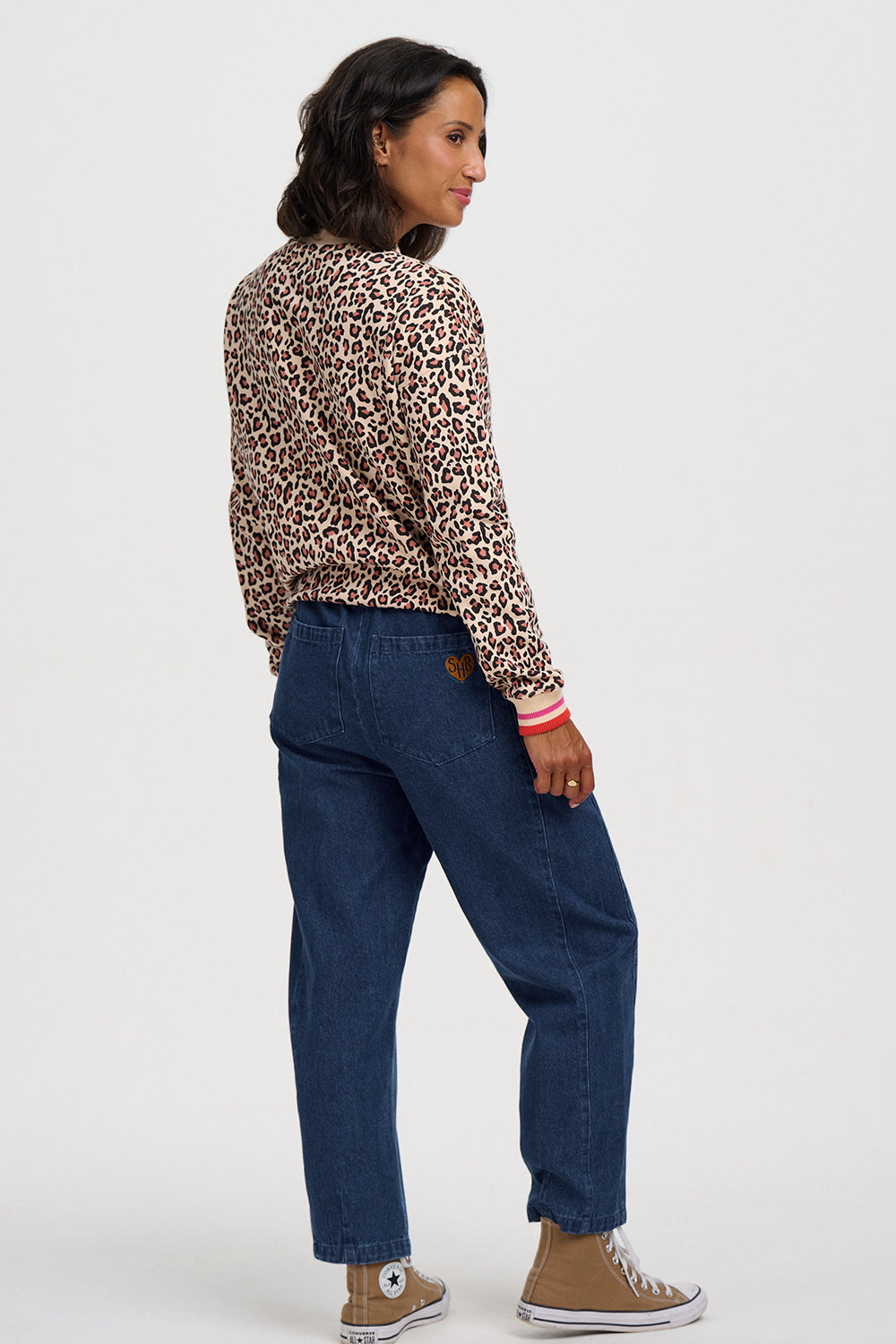 A woman shown from the back wearing a leopard-print long-sleeve top, dark blue jeans, and tan sneakers against a plain light gray studio background.