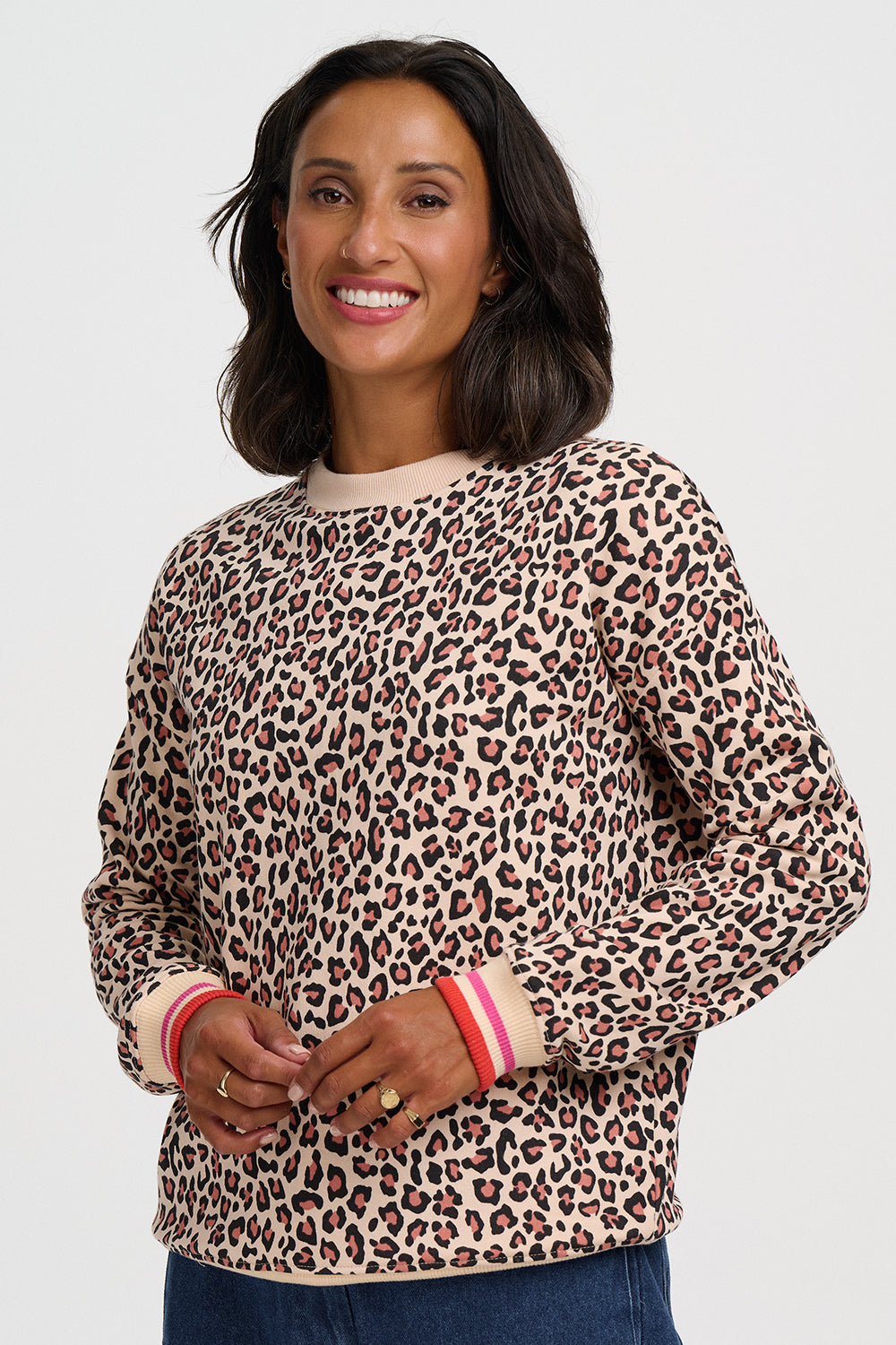 A woman wearing a beige crewneck sweatshirt with a brown and black leopard print pattern, featuring pink and white striped ribbed cuffs, set against a plain light gray background.