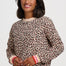 A woman wearing a beige crewneck sweatshirt with a brown and black leopard print pattern, featuring pink and white striped ribbed cuffs, set against a plain light gray background.