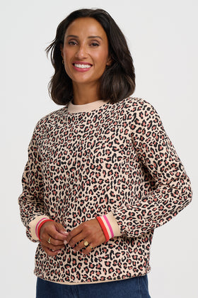 A woman wearing a beige crewneck sweatshirt with a brown and black leopard print pattern, featuring pink and white striped ribbed cuffs, set against a plain light gray background.