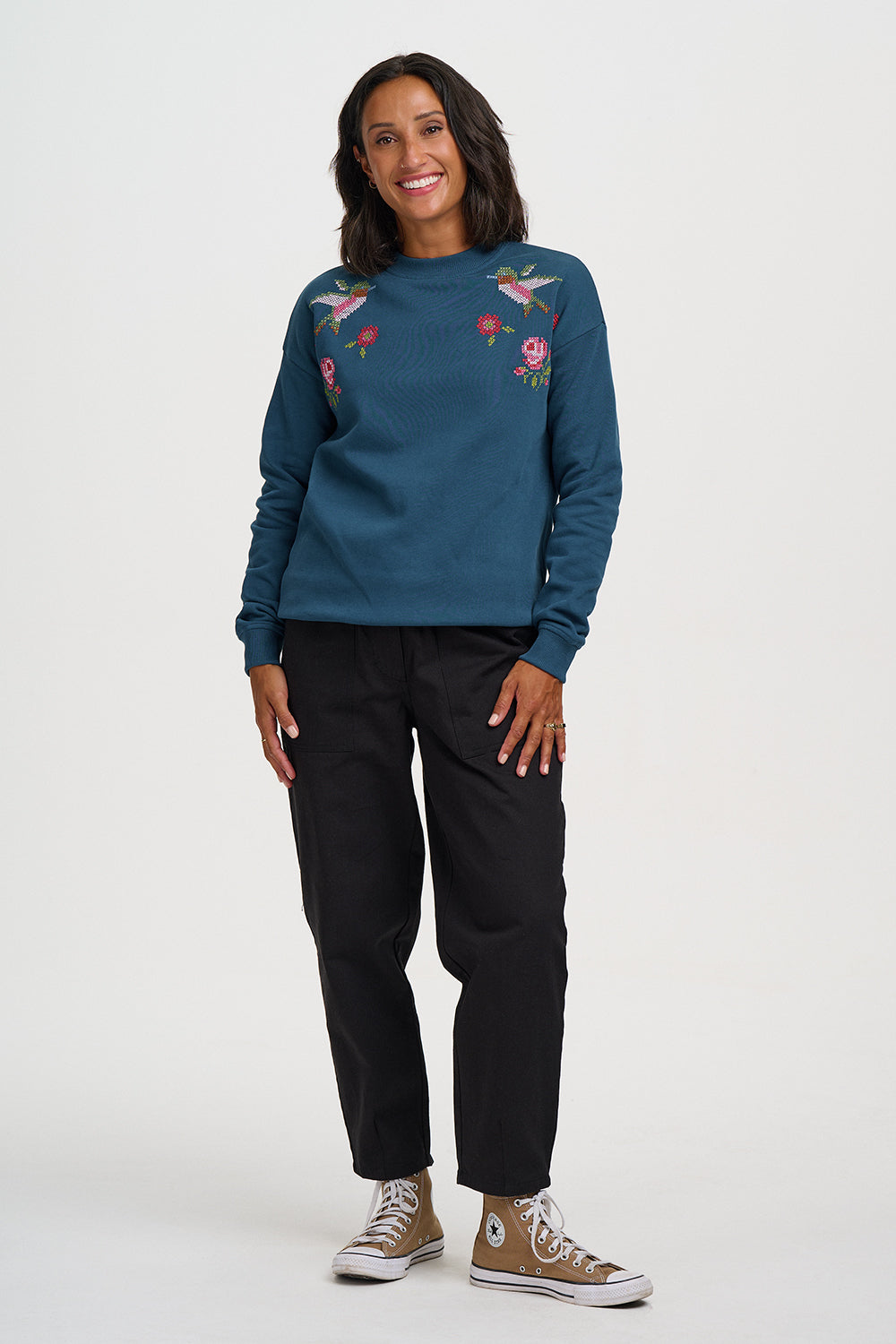 A woman modeling a teal crewneck sweatshirt with colorful floral and bird embroidery on the chest, paired with black pants and brown Converse sneakers, against a plain white studio background.