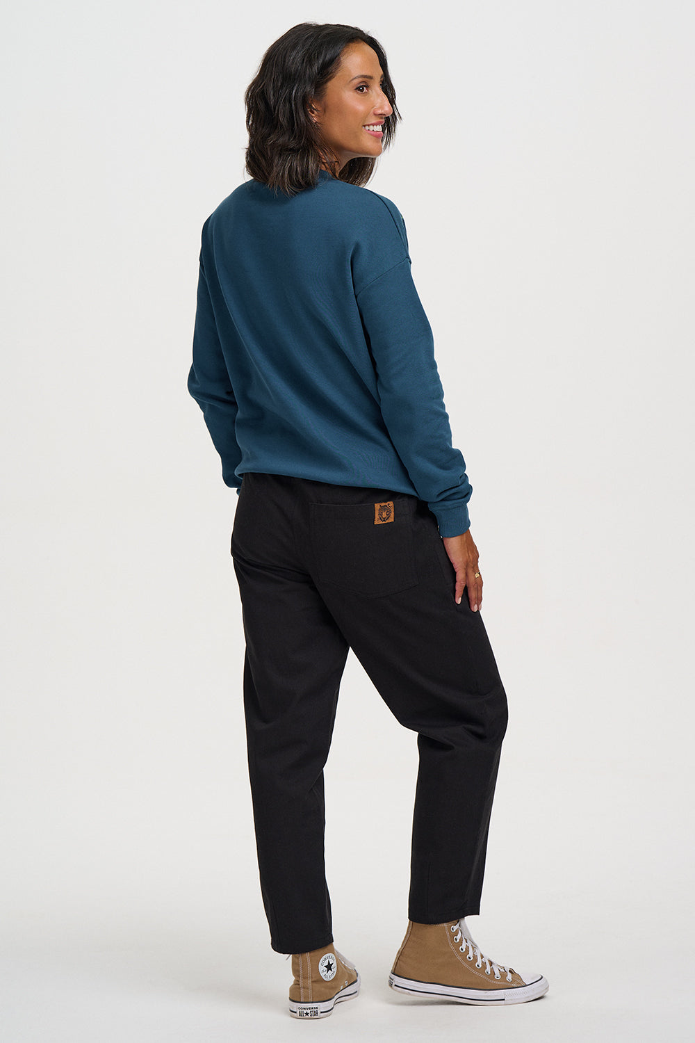 A woman shown from the back wearing a dark teal long-sleeve crewneck sweatshirt, black pants, and tan Converse high-top sneakers against a plain white studio background.