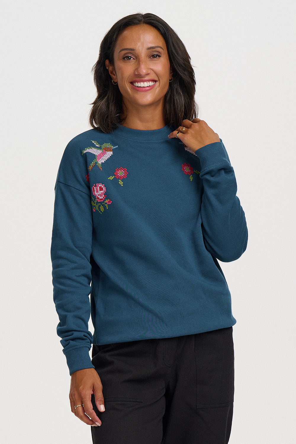 A woman modeling a teal blue long-sleeve crewneck sweatshirt with colorful embroidered flowers and a bird on the chest, paired with black pants, against a light gray studio background.
