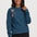 A woman modeling a teal blue long-sleeve crewneck sweatshirt with colorful embroidered flowers and a bird on the chest, paired with black pants, against a light gray studio background.