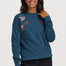 A woman modeling a teal blue long-sleeve crewneck sweatshirt with colorful embroidered flowers and a bird on the chest, paired with black pants, against a light gray studio background.