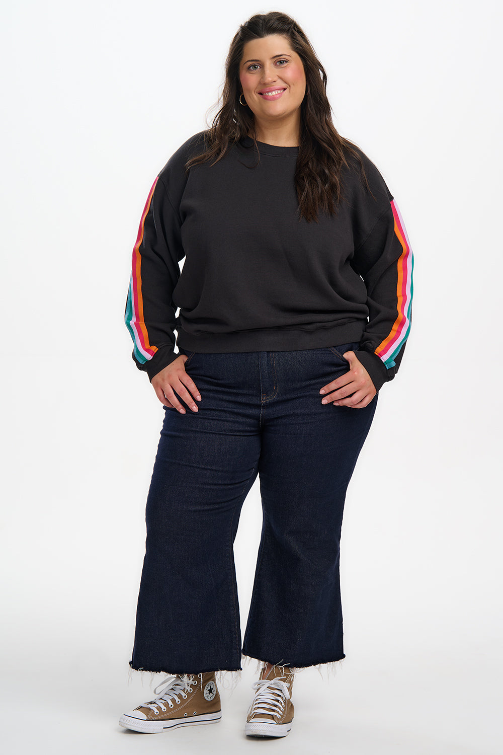 Person wearing a black sweatshirt with rainbow sleeves and dark jeans on a white background