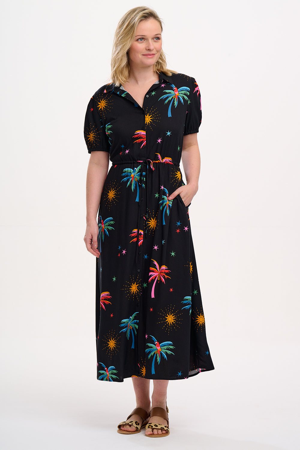 A model wearing a black maxi shirt dress with a palm tree and sun print. The dress features a button-front design, puff sleeves, a drawstring waist, and pockets.
