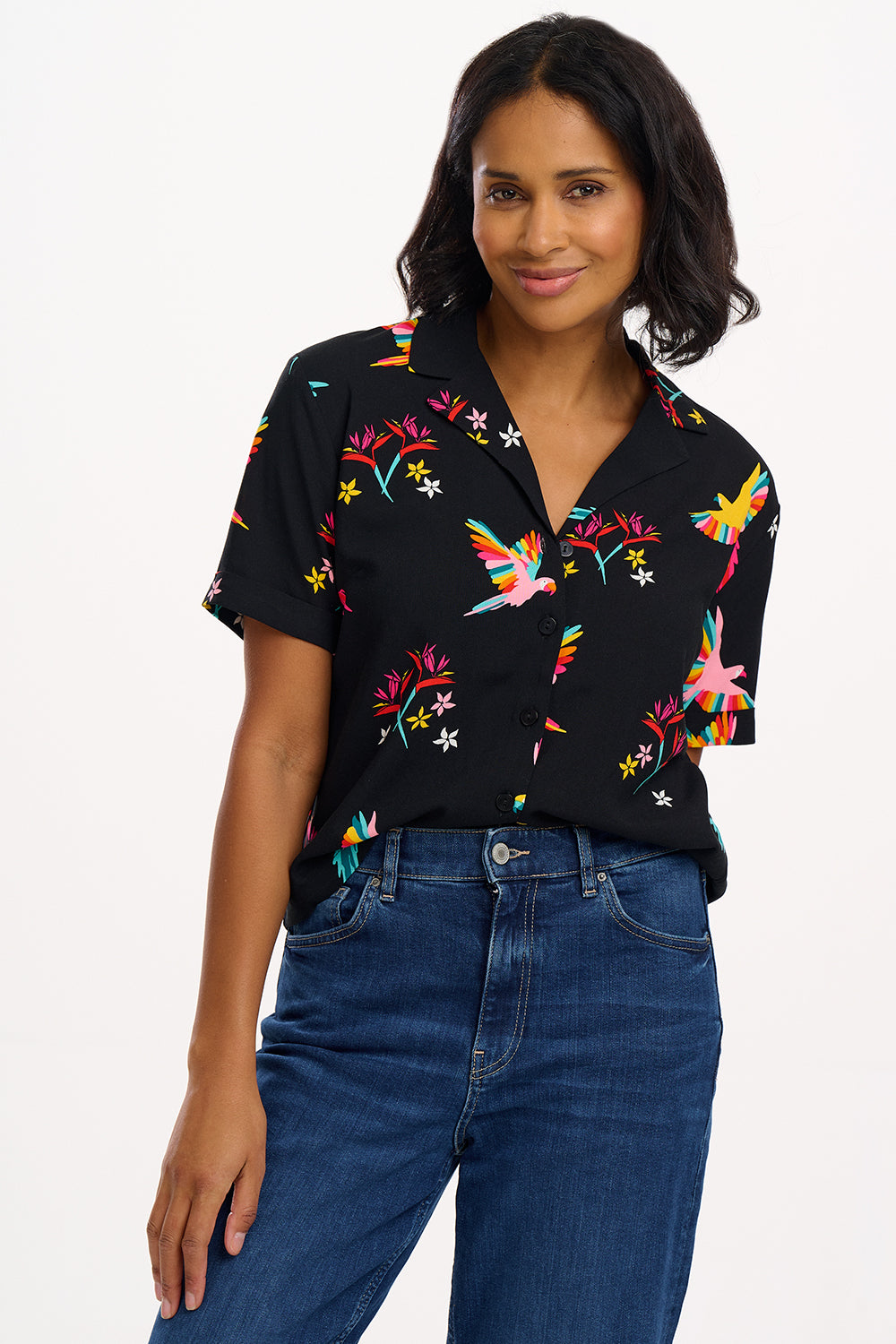 A woman smiling and wearing a short-sleeved black shirt with a rainbow parrots and flowers print, paired with blue jeans.