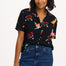 A woman smiling and wearing a short-sleeved black shirt with a rainbow parrots and flowers print, paired with blue jeans.