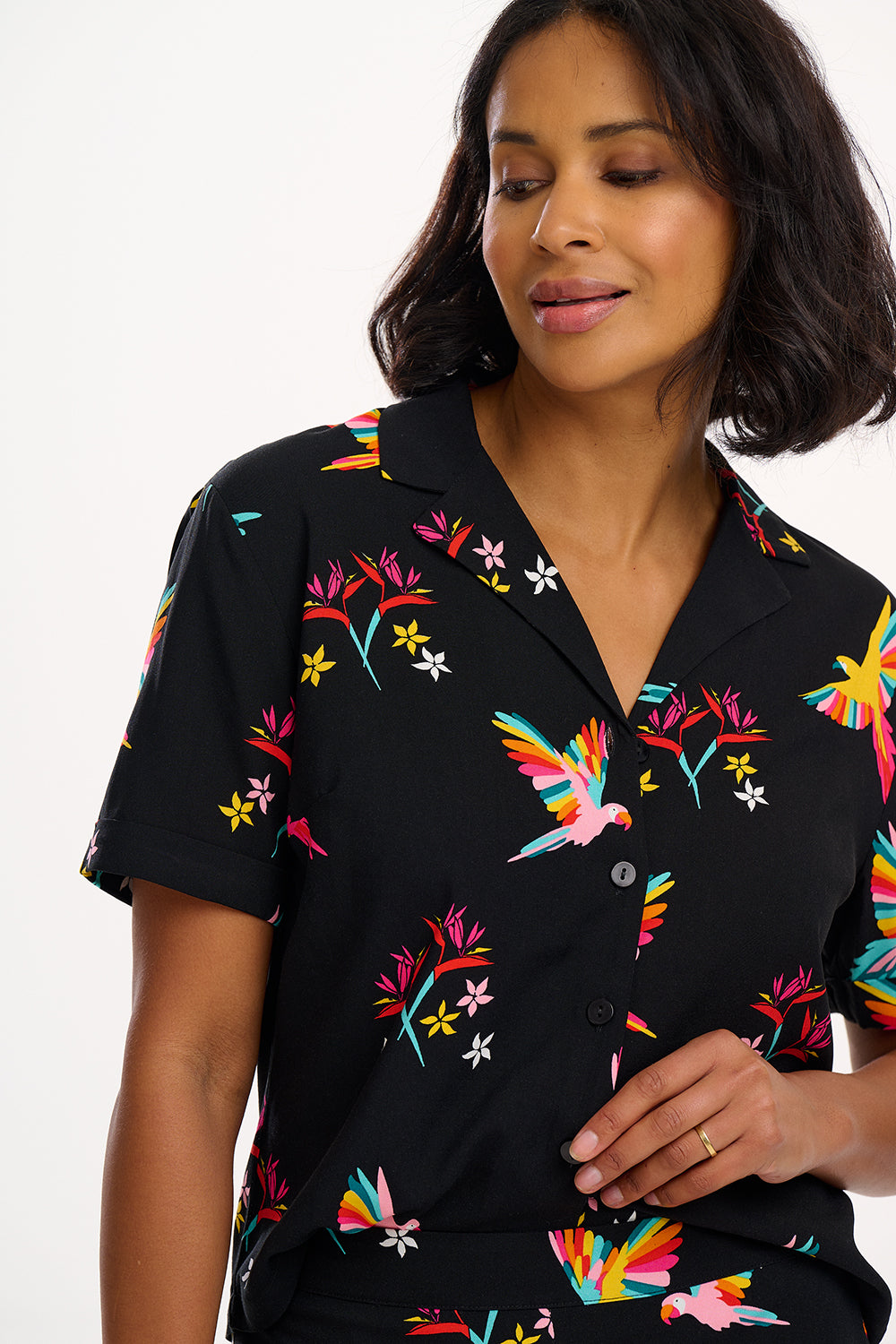 A woman smiling and wearing a short-sleeved black shirt with a rainbow parrots and flowers print, paired with blue jeans.