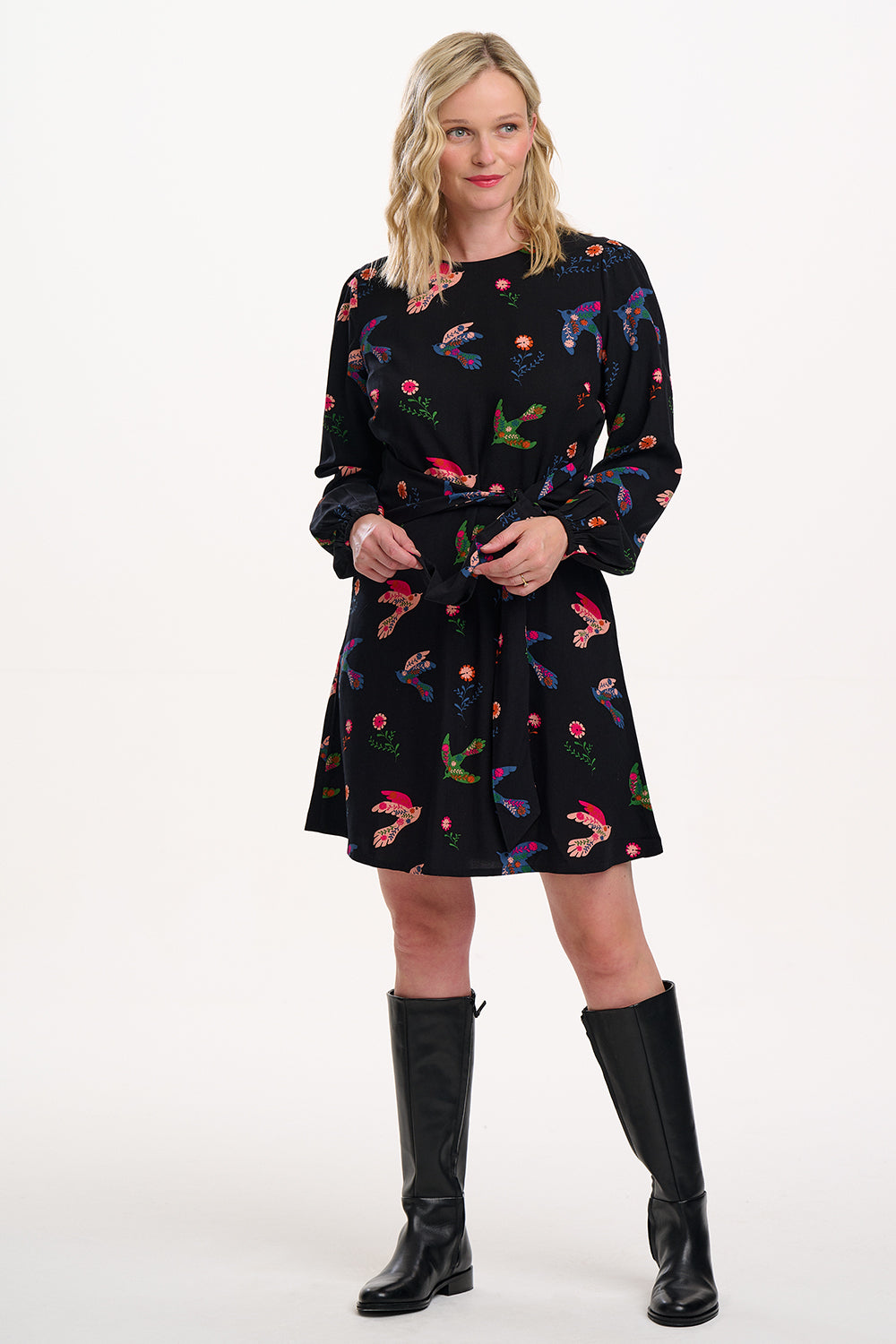 Sugarhill brighton model wearing a black dress with colourful floral pattern and black knee-high boots on a white background
