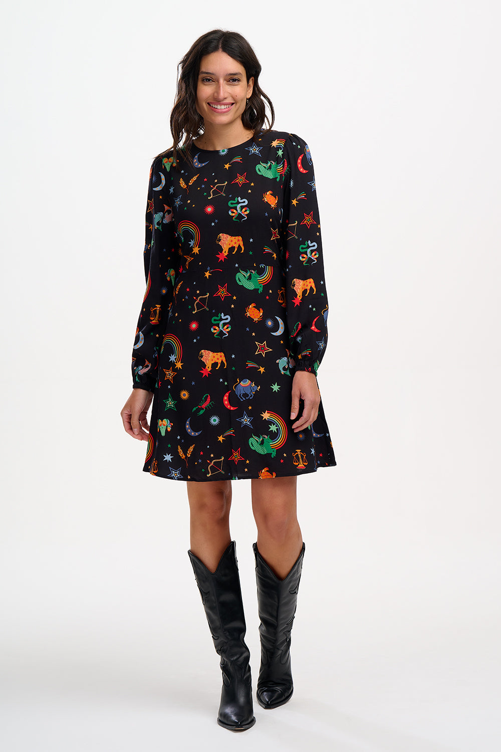 A woman standing and wearing a black mini dress with a Zodiac Rainbow Skies print featuring multicolored celestial icons on a black base.