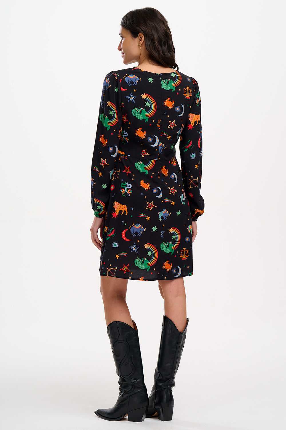 A woman standing and wearing a black mini dress with a Zodiac Rainbow Skies print featuring multicolored celestial icons on a black base.