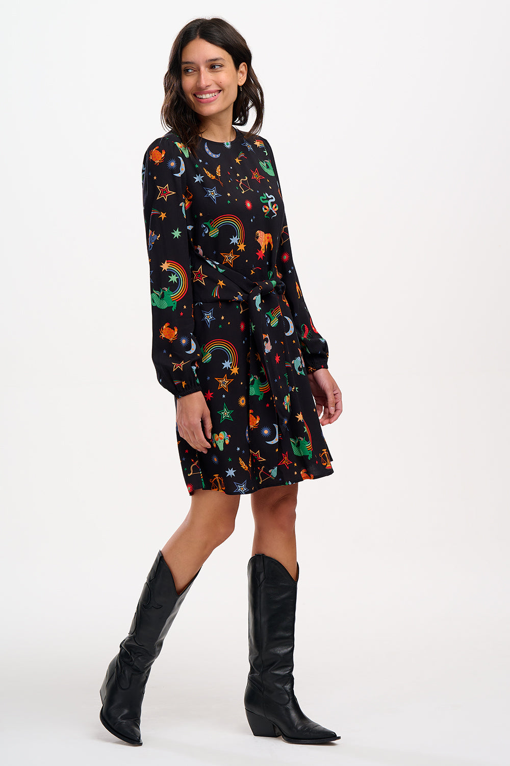 A woman standing and wearing a black mini dress with a Zodiac Rainbow Skies print featuring multicolored celestial icons on a black base.