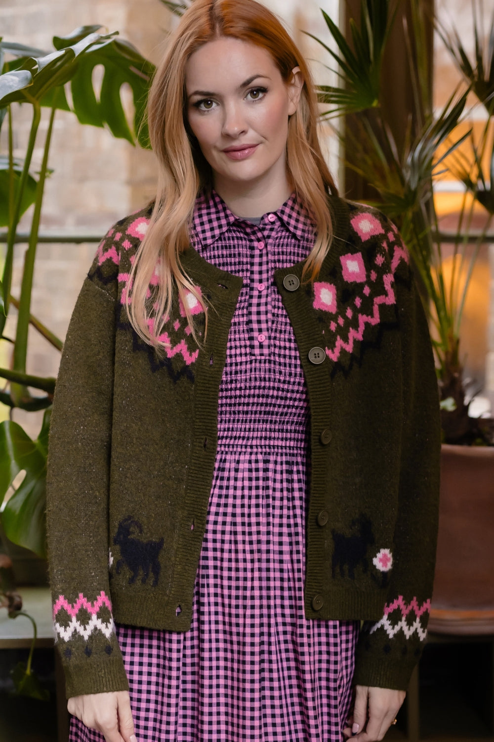 A woman wearing an olive green knit cardigan with pink and white geometric patterns and black dog motifs, layered over a pink and purple gingham dress, posed in front of a warm indoor background with plants.