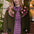 A woman wearing an olive green knit cardigan with pink and white geometric patterns and black dog motifs, layered over a pink and purple gingham dress, posed in front of a warm indoor background with plants.