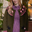 A woman wearing an olive green knit cardigan with pink and white geometric patterns and black dog motifs, layered over a pink and purple gingham dress, posed in front of a warm indoor background with plants.