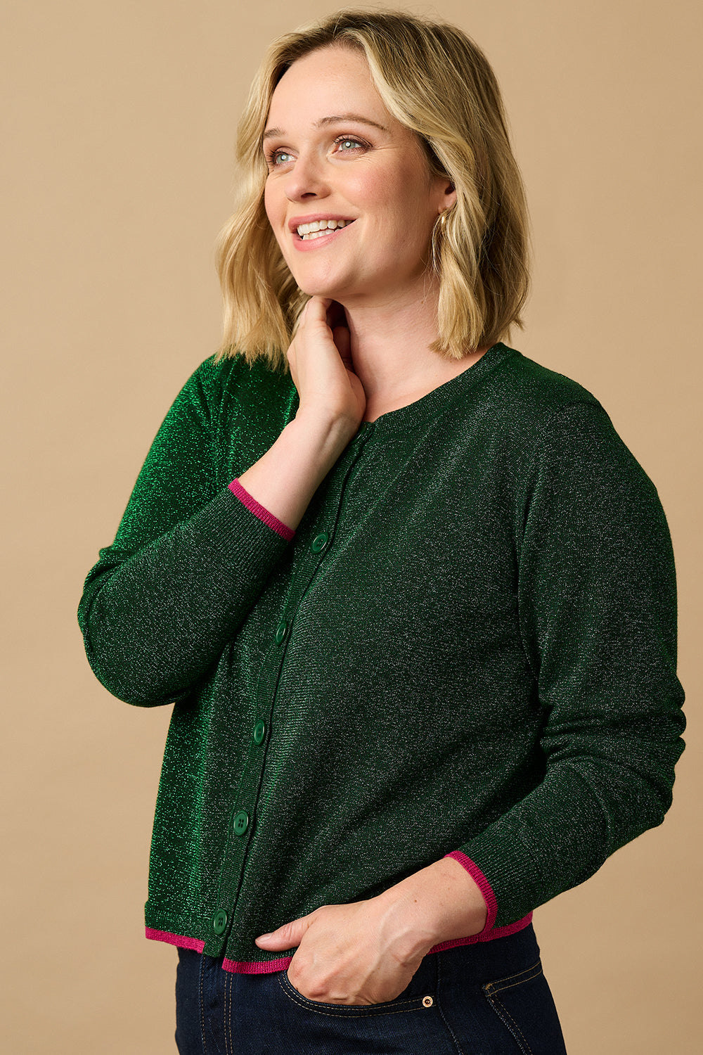 A woman wearing a green cardigan with metallic fibers and pink stripes, featuring a deep V-neckline and ribbed cuffs. She is also wearing a pink shirt and blue jeans.