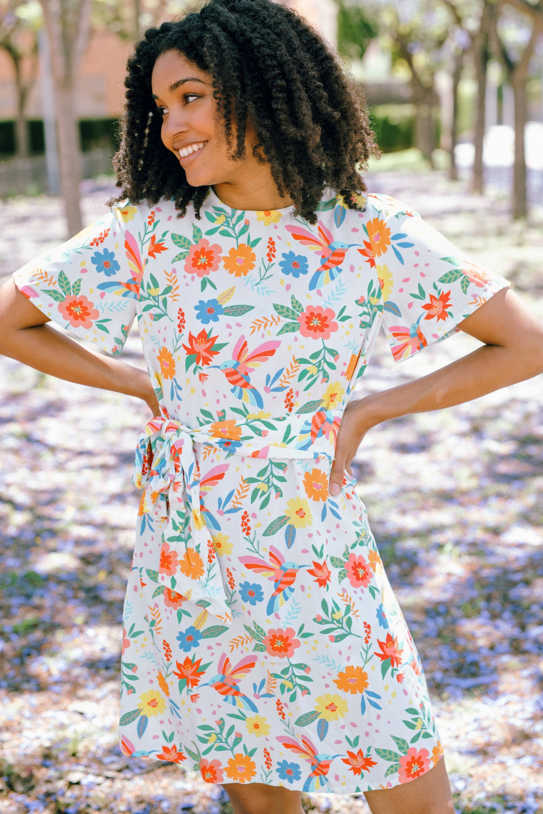 A woman wearing an off-white mini dress with a tropical hummingbird print, featuring a rounded neckline and fluted sleeves.
