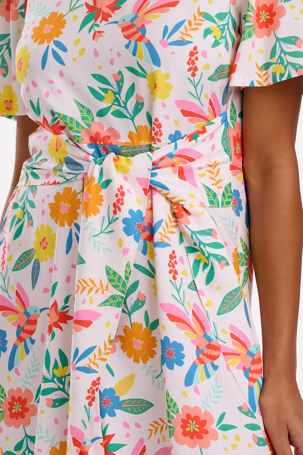 A woman wearing an off-white mini dress with a Tropical Hummingbird print, featuring fluted sleeves and a self-tie belt.