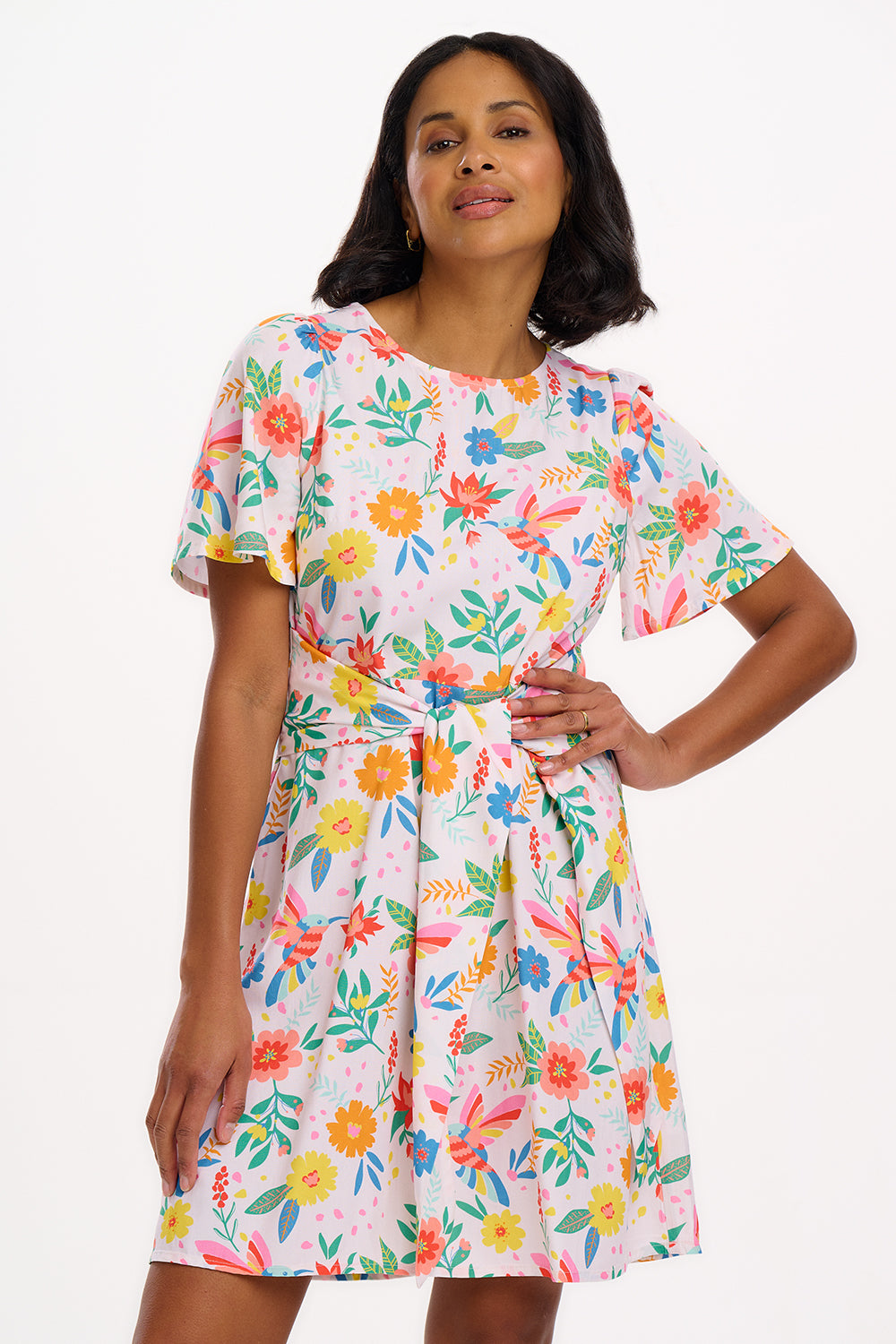 A woman wearing an off-white mini dress with a Tropical Hummingbird print, featuring fluted sleeves and a self-tie belt.