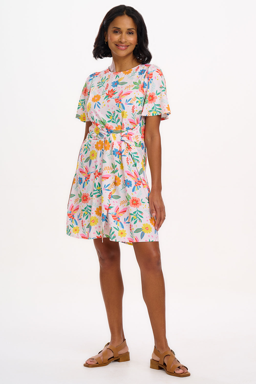 A woman wearing an off-white mini dress with a Tropical Hummingbird print, featuring fluted sleeves and a self-tie belt.