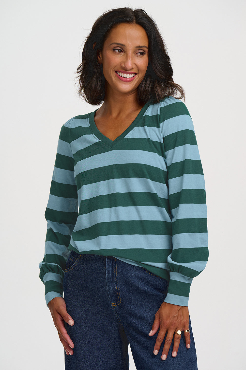 A woman wearing a long-sleeve V-neck top with horizontal dark green and light blue stripes, paired with dark blue jeans, against a plain light gray background.