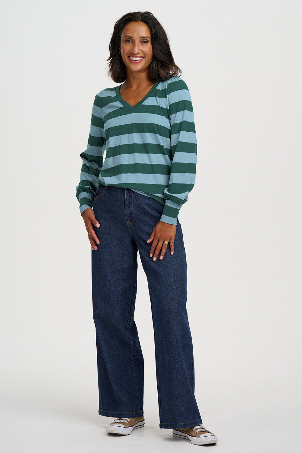A woman modeling a green and light blue striped long-sleeve V-neck top with dark blue wide-leg jeans and brown sneakers, standing against a light gray studio background.